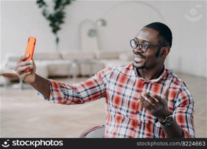 Modern african american man wearing glasses holding smartphone talking online by video call. Friendly smiling young black guy blogger communicates in social networks, looking at phone screen.. Modern african american man blogger in glasses holding smartphone talking online by video call