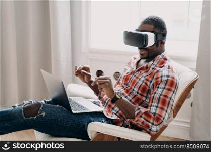 Modern african american man wearing vr glasses working in cyberspace at laptop, black guy developer or designer interacting with virtual reality objects, sitting in armchair. Distant work and tech.. African american man in vr glasses working in cyberspace at laptop, interacting with virtual reality
