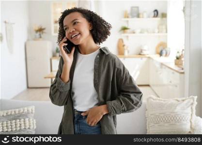 Modern african american young female answer call, talking on phone with a friend at home on sunny day. Biracial teen girl enjoying conversation by smartphone discussing positive good news on weekend.. Biracial girl answer call, talking on phone with friend, discussing good news at home on sunny day