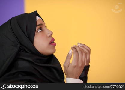 Modern African Muslim woman makes traditional prayer to God, keeps hands in praying gesture, wears traditional white clothes, has serious facial expression, isolated over plastic yellow background
