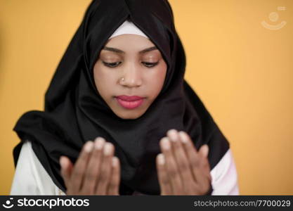 Modern African Muslim woman makes traditional prayer to God, keeps hands in praying gesture, wears traditional white clothes, has serious facial expression, isolated over plastic yellow background
