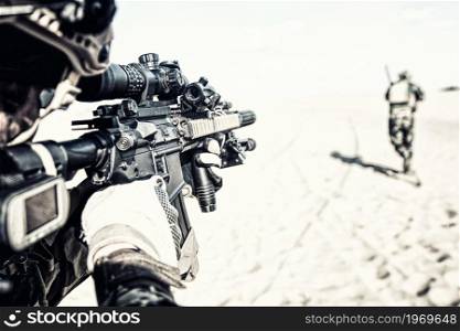 Modern army soldier aiming through service rifle optical sight while running after teammate in hot sandy area or dessert. Special forces infantry covering brother in arms in action. Over shoulder view. Commando aiming gun ana running in desert area