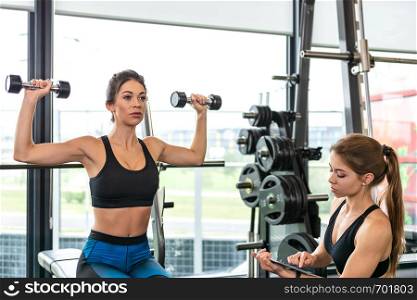 Modern athletic workout concept - fitness woman is training with dumbbells at the gym and is leading by a trainer and an application on a tablet.