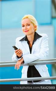 Modern business woman with cell phone leaning on railing at office building&#xA;