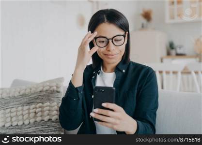 Modern businesswoman uses mobile apps on smartphone straightening glasses, sitting on sofa at home. Female holding phone, shopping in online store, checking social networks news on couch. E-commerce. Modern businesswoman uses mobile apps on smartphone straightening glasses, sitting on sofa at home