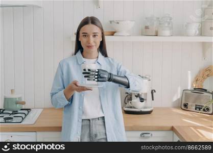 Modern caucasian disabled girl holding cup of coffee, using bionic prosthetic arm, standing in kitchen. Young woman holds mug by artificial hand at home. Lifestyle of people with disabilities concept.. Disabled girl holding cup by bionic prosthetic arm in kitchen. Lifestyle of people with disabilities