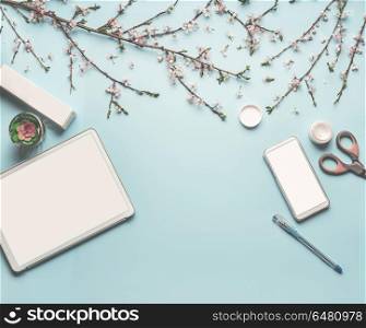 Modern desktop workplace flat lay with mock up of tablet computer and smart phone, cosmetic products, spring blossom branches , scissors, pen and green leaves on blue background, top view, frame
