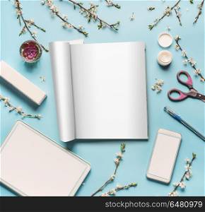 Modern desktop workplace flat lay with mock up open magazine , tablet computer and smart phone, cosmetic products, spring blossom, scissors, pen and green leaves on blue background, top view.