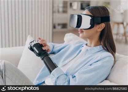 Modern disabled girl in VR glasses interacts with augmented reality, touching her high tech bionic prosthetic arm, sitting on sofa at home. Disability and medical future technologies concept.. Disabled girl in VR glasses interacts with augmented reality, touches her high tech prosthetic arm