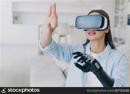 Modern disabled woman wearing virtual reality glasses experiencing augmented reality apps, using bionic prosthetic arm. Girl with artificial robotic hand in VR goggles, watching 360 degrees video.. Disabled girl in virtual reality glasses experiencing augmented reality, using bionic prosthetic arm