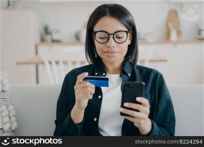Modern female holding credit card and smartphone uses mobile bank app, online banking services. Focused woman pays for purchases, shopping on internet, sitting on couch at home. E-banking.. Modern female holding credit card, phone, uses mobile bank app, online banking services at home