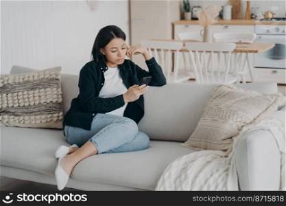 Modern female uses mobile apps at home spending leisure time online with smartphone sitting on couch. Focused young woman answers messages in social networks, browsing news, shopping on internet.. Modern female uses mobile apps at home spending leisure time online with smartphone sitting on couch