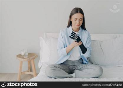 Modern girl with disability training to install her bionic prosthetic arm, sitting on bed at home. Young woman using high tech prosthesis after limb loss. Advertising of robotic artificial limbs.. Modern girl with disability training to install her bionic prosthetic arm, sitting on bed at home