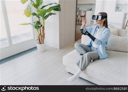 Modern girl with disability, wearing vr glasses moving artificial hand, sitting on sofa. Disabled young woman gets rehabilitation using sensory bionic prosthetic arm, interacting with virtual reality.. Girl with disability in vr glasses interacts with virtual reality by bionic prosthetic arm at home