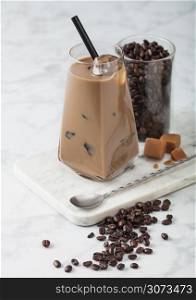 Modern glass of iced coffee with milk on marble board with jar of coffee beans and salted caramel and long spoon on light table background.