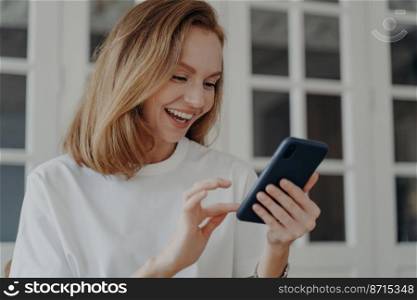 Modern happy woman using mobile apps, holding phone in hands, chatting in social networks. Smiling young female uses smartphone, reading good news, online shopping. E-commerce concept.. Happy young woman using mobile apps, holding modern smartphone, shopping online. E-commerce concept