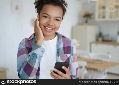Modern happy young biracial teen girl in wireless earphones holding smartphone listens to music, radio song. Smiling mixed race female enjoying melody sound, using music apps at home.. Modern happy young biracial girl holding smartphone listens to music by earphone, uses musical apps