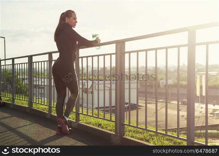Modern healthy lifestyle. A woman in sportswear with bottle of water have rest at sunset on the evening city background