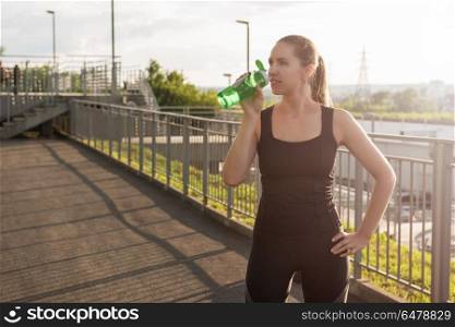 Modern healthy lifestyle. A woman in sportswear with bottle of water have rest at sunset on the evening city background