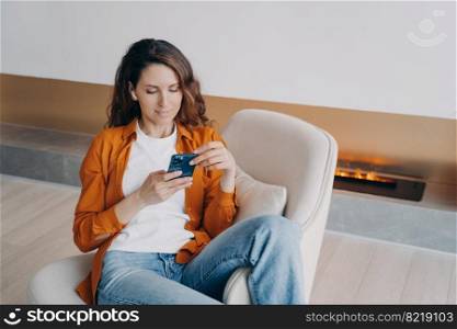 Modern hispanic woman in wireless earphones using smartphone apps, choosing music playlist, audio book, sitting in armchair. Female browsing social networks, shopping online, listening to new podcast.. Modern girl in earphones using smartphone apps, shopping online, listening to new podcast or music