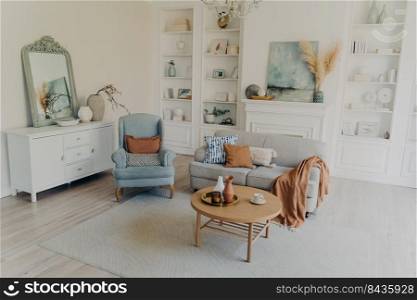 Modern home interior. Pillows on sofa and armchair. Mirror over chest. Decoration with pots and vases on shelves. Table near couch. Elegant scandinavian interior, cozy hotel room concept.. Modern home or cozy hotel room. Sofa and armchair, Mirror over chest, decoration and textile.