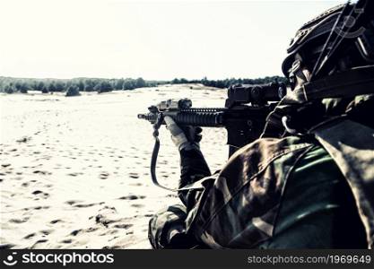 Modern infantryman lying on ground and aiming assault rifle with optical sight. Army special operations fighter visually inspecting area, monitoring movement of enemy in sandy area. Over shoulder view. Marine raider aiming service rifle in sandy area