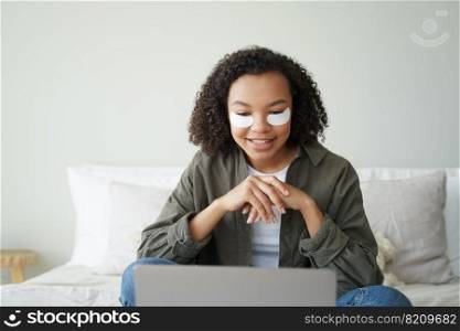 Modern mixed race girl with under eye patches on face working at laptop in bedroom. Biracial young lady with under-eye patch watching series on computer, sitting on bed. Skincare beauty routine.. Mixed race girl with under eye patches on face working at laptop in bedroom. Skincare beauty routine
