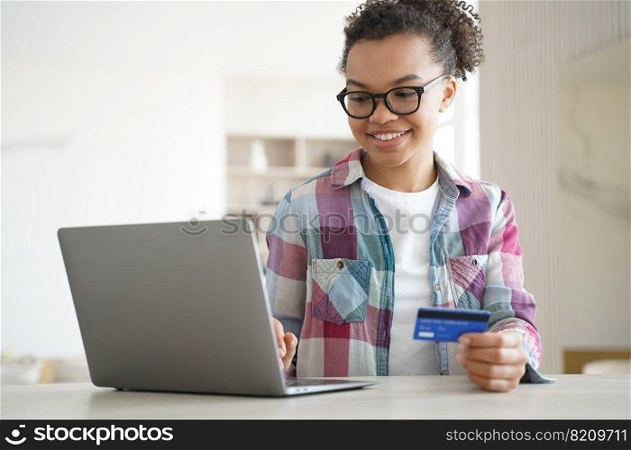 Modern mixed race teen girl holds bank credit card uses online banking services on laptop. Smiling young biracial lady paying shopping on internet on computer at home. E-banking, e-commerce concept.. Mixed race teen girl holds bank credit card uses online banking services on laptop. E-banking