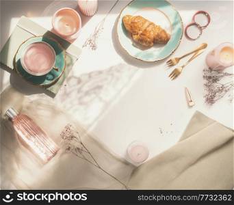 Modern morning routine with croissant, flowers tea, scented candles, forks, pink perfume and notebook on white table. Romantic feminine background. Top view with copy space.
