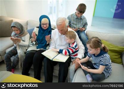 modern muslim family grandparents with grandchildren reading Quran and praying together on the sofa before iftar dinner during a ramadan feast at home