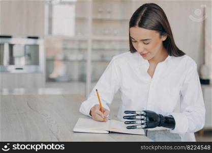 Modern pretty girl with disability writing in notebook, sitting at table at home. Disabled young female makes notes, writes to-do list, planning goals in notepad, using bionic prosthetic arm.. Young girl with disability writing to-do list in notebook planning goals using bionic prosthetic arm
