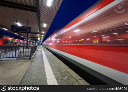 Modern railway station with high speed passenger train on railroad track in motion at night in Nuremberg, Germany. Fast red commuter train.. Industrial landscape