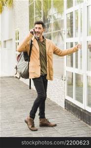 modern smiling man with his backpack talking mobile phone while opening glass door
