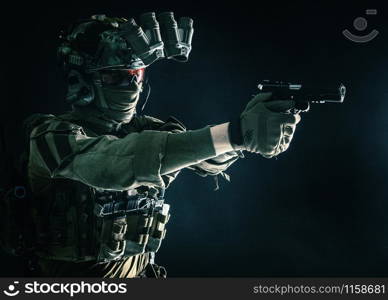 Modern soldier, counter terrorist squad fighter in combat uniform, helmet and tactical radio headset, aiming service pistol in darkness, shooting with handgun, studio portrait on dark background. Counter terrorist squad fighter soldier aiming pistol