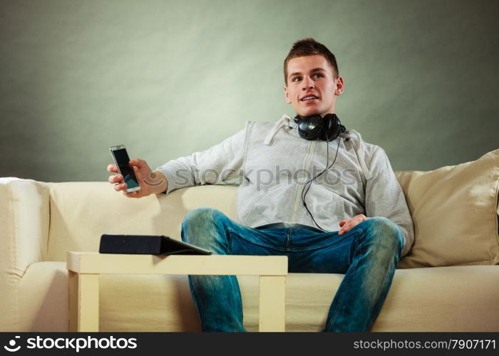 Modern technologies connection leisure concept. Young handsome man relaxing on couch with headphones smartphone and tablet at home