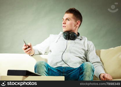 Modern technologies connection leisure concept. Young handsome man relaxing on couch with headphones smartphone and tablet at home