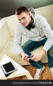 Modern technologies connection leisure concept. Young man relaxing on couch with headphones smartphone and tablet at home unusual view