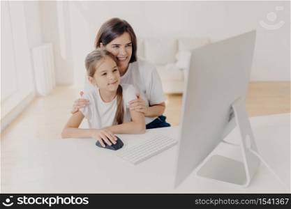 Modern technology and family concept. Caring lovely mother teaches little daughter how to use internet, explains how to download application, pose in spacious room with white walls, spend time at home