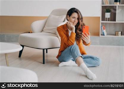 Modern woman, holding smartphone, taking selfie photo or chatting by video call, sitting on heated floor in living room. Female wearing earphones communicates online using mobile phone app at home.. Modern girl holding smartphone taking selfie photo or chatting by video call sitting on heated floor