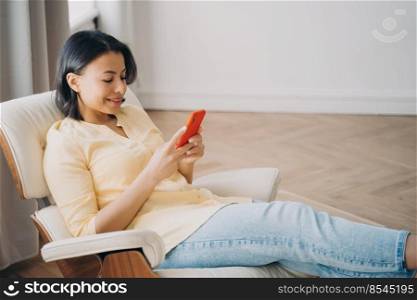 Modern woman using phone, sitting in comfortable armchair at home. Female looking at smartphone screen, chatting messaging or shopping in online store. E-commerce, mobile apps advertising.. Female using phone sitting in comfortable armchair at home, shopping online. Mobile apps advertising