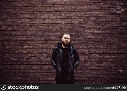 Modern young bearded man in black style clothes posing against brick wall.. Modern young bearded man in black style clothes posing against brick wall