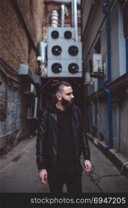 Modern young bearded man in black style clothes standing in urban background.. Modern young bearded man in black style clothes standing in urban background