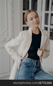 Modern young businesswoman wearing casual clothing posing indoors. Pretty female model in white suit jacket and blue jeans poses for portrait, looking at camera. Women&rsquo;s fashion concept.. Modern pretty young businesswoman wearing casual clothing posing for portrait. Women&rsquo;s fashion