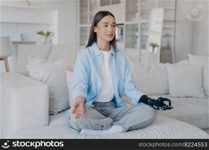 Modern young disabled girl makes mudra gesture with bionic prosthetic arm sitting in lotus posture on sofa. Female with artificial hand practicing yoga at home. Disability and healthy lifestyle.. Young disabled girl with bionic prosthetic arm practices yoga at home. Disability, healthy lifestyle