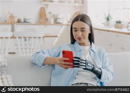 Modern young disabled girl with bionic prosthetic arm chatting in social networks, holding phone with artificial robotic hand, sitting on sofa at home. Disability, bionic prosthesis advertising.. Disabled girl with bionic prosthetic arm holds smartphone. Disability, bionic prosthesis advertising