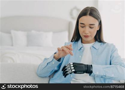 Modern young girl with artificial limb trains to use her bionic prosthetic hand, sitting on sofa at home. Pretty disabled woman moving, raising arms. Lifestyle of people with disabilities.. Modern young girl with artificial arm trains to use bionic prosthetic hand, sitting on sofa at home