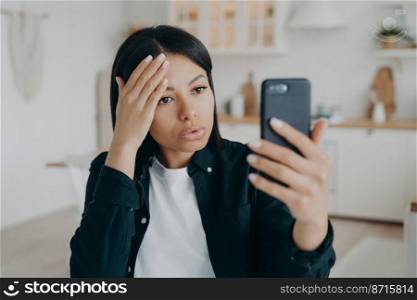Modern young woman blogger holding smartphone, makes video call or takes a selfie photo, standing at home. Focused female has online communication on cellphone, using mobile phone camera.. Modern young woman blogger holding smartphone, makes video call or takes a selfie photo at home
