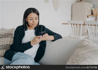 Modern young woman uses mobile apps holding smartphone, shopping online at home. Female texting message on phone, chatting in social networks, sitting on comfortable couch. E-commerce.. Female uses mobile apps holding smartphone, chatting in social networks, shopping online at home