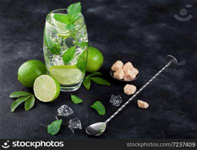Mojito refreshing summer cocktail in glass with ice cubes mint and lime on black board with spoon and fresh limes with cane sugar.