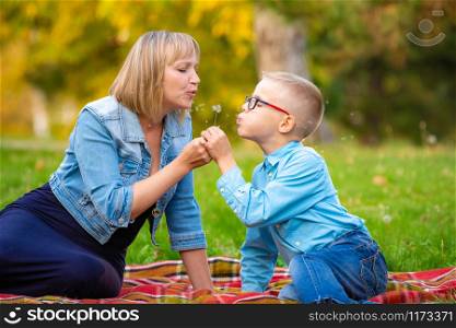 Mom and boy teen sit on green glade and blow off dandelion